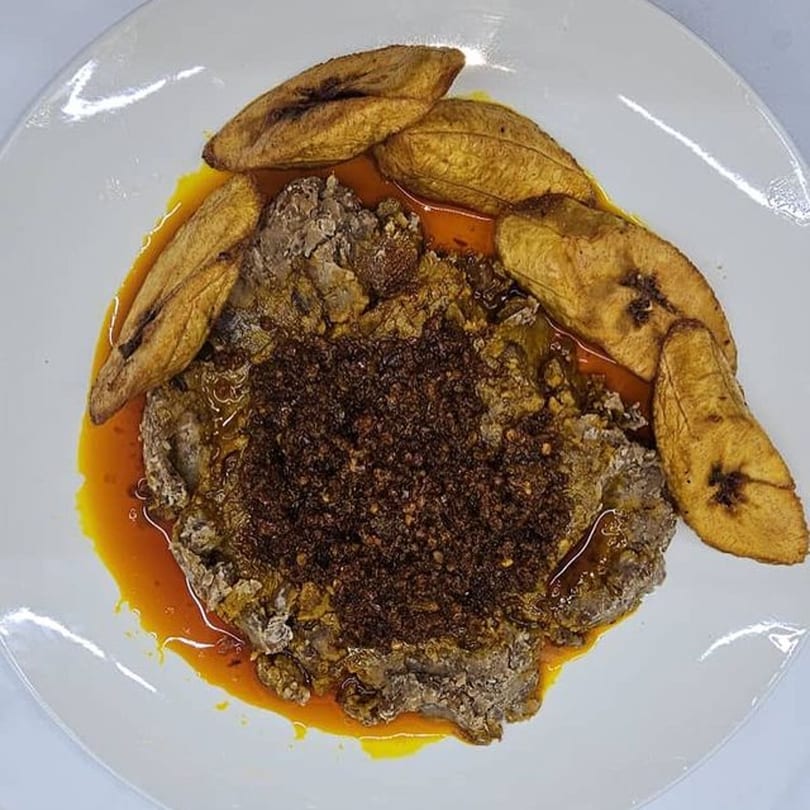 African dish with dark meat, a rich sauce, and fried plantains on a white plate.