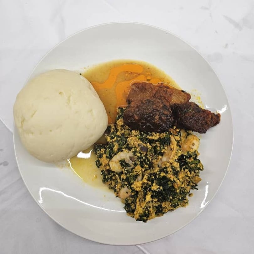 A white plate holds a mound of fufu, stewed greens with shrimp, and dark meat in sauce.